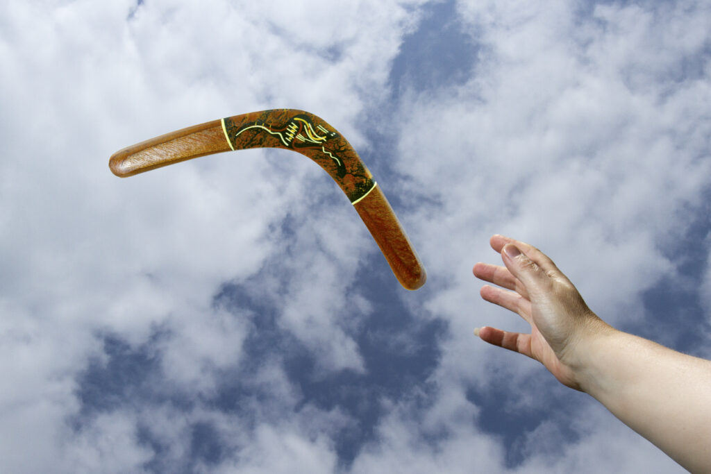 Hand catching a painted, wooden boomerang midair with blue sky and cloud background.